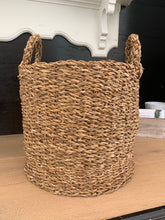Load image into Gallery viewer, A brown woven basket in front of a black wall and a white shelf.
